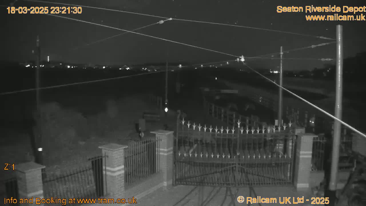 A nighttime, black-and-white security camera view shows an open gate with a fence, several power lines overhead, and a dimly lit area beyond.