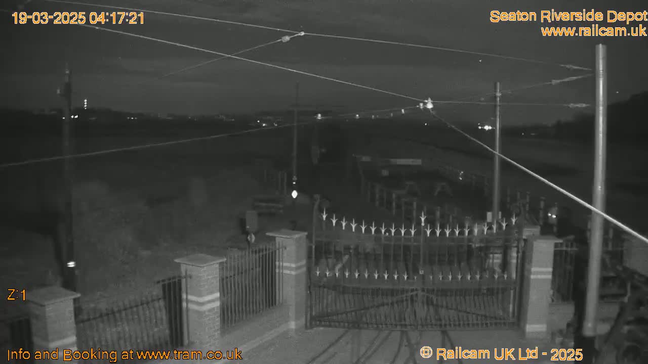 A nighttime, low-light view of a gated entrance to what appears to be a transit depot, with overhead power lines visible.