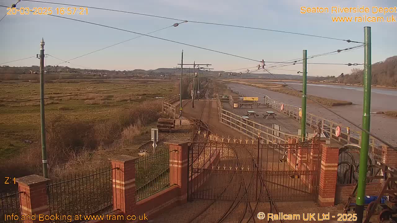 A sunny view of a train depot with railway tracks and gates leading to a river under a clear sky.