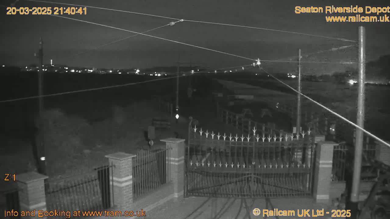 A nighttime, black and white image shows a closed gate at the entrance to a depot, with overhead wires and a distant city skyline faintly visible under a dark sky.