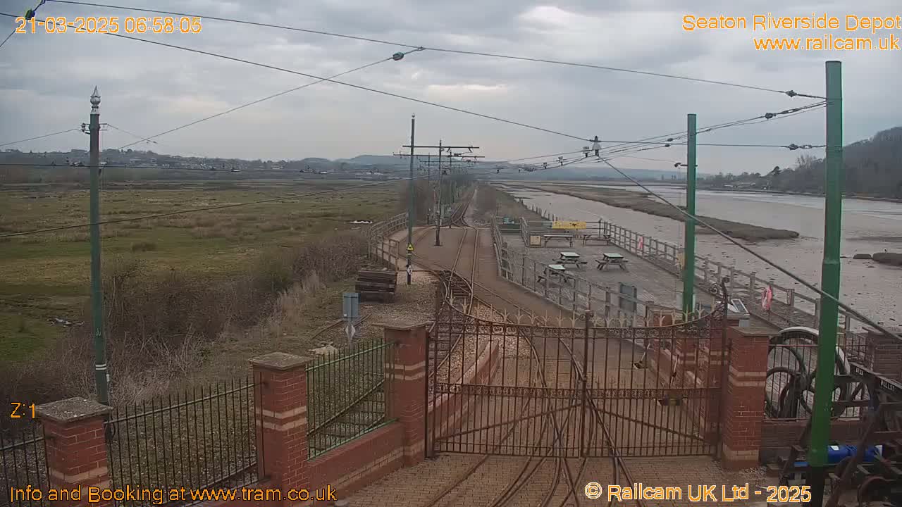 A view of train tracks leading to a depot beside a calm, muddy river under an overcast sky.