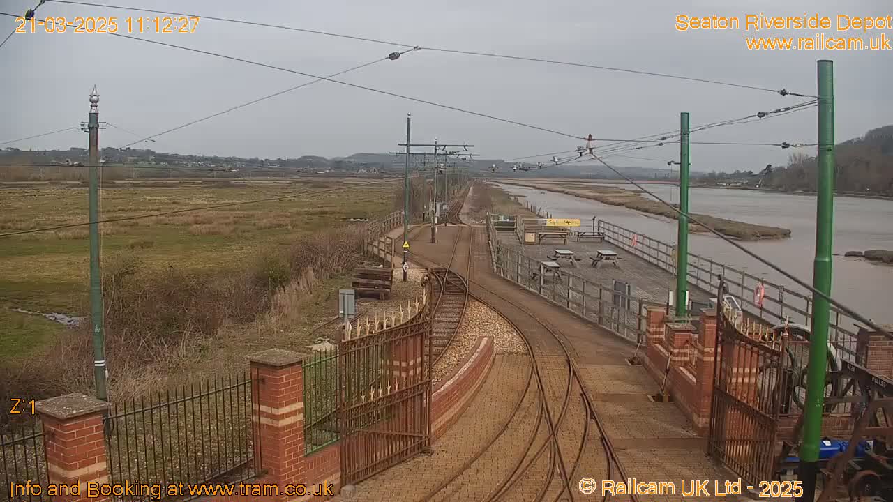 A railway track curves alongside a river under an overcast sky, passing by a fenced-in depot area with picnic tables.