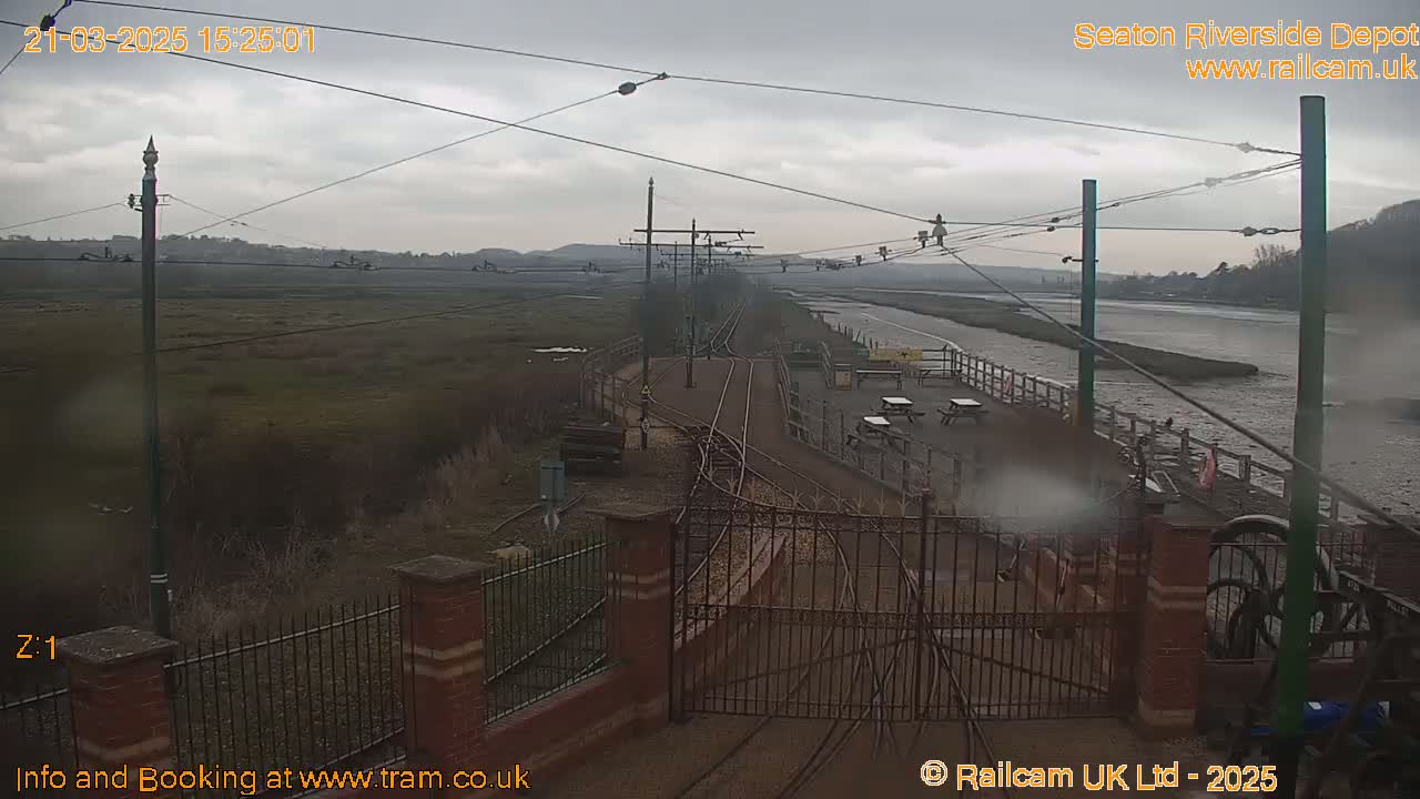 An overcast day shows a train depot alongside a river, featuring train tracks, a gate, and picnic tables.