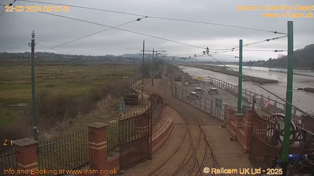 A railway depot with tracks curving towards a river under an overcast sky.