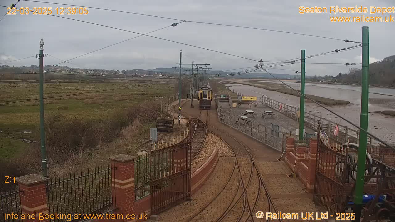 A yellow tram travels along train tracks toward a depot, next to a river under an overcast sky.