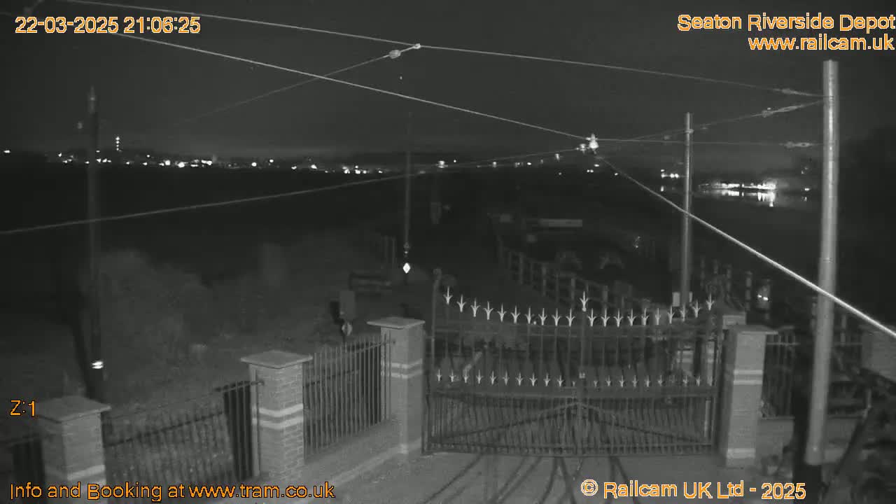 A nighttime, black-and-white security camera view shows an open gate at a depot with overhead wires and distant lights visible in the background.