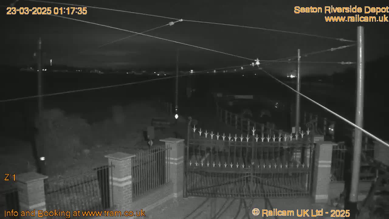 A nighttime, low-light view of a gated area with overhead wires and distant lights.