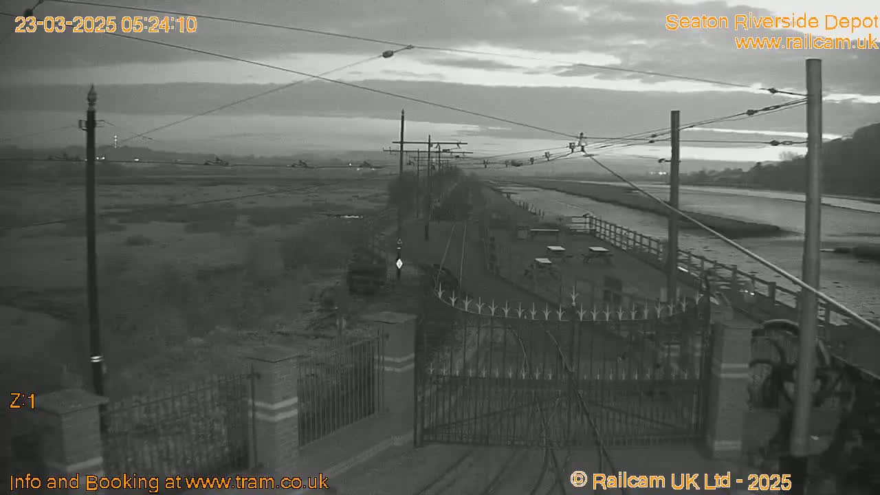 A grayscale, pre-dawn view of a waterfront depot with overhead wires, a gate, and a calm body of water beyond.