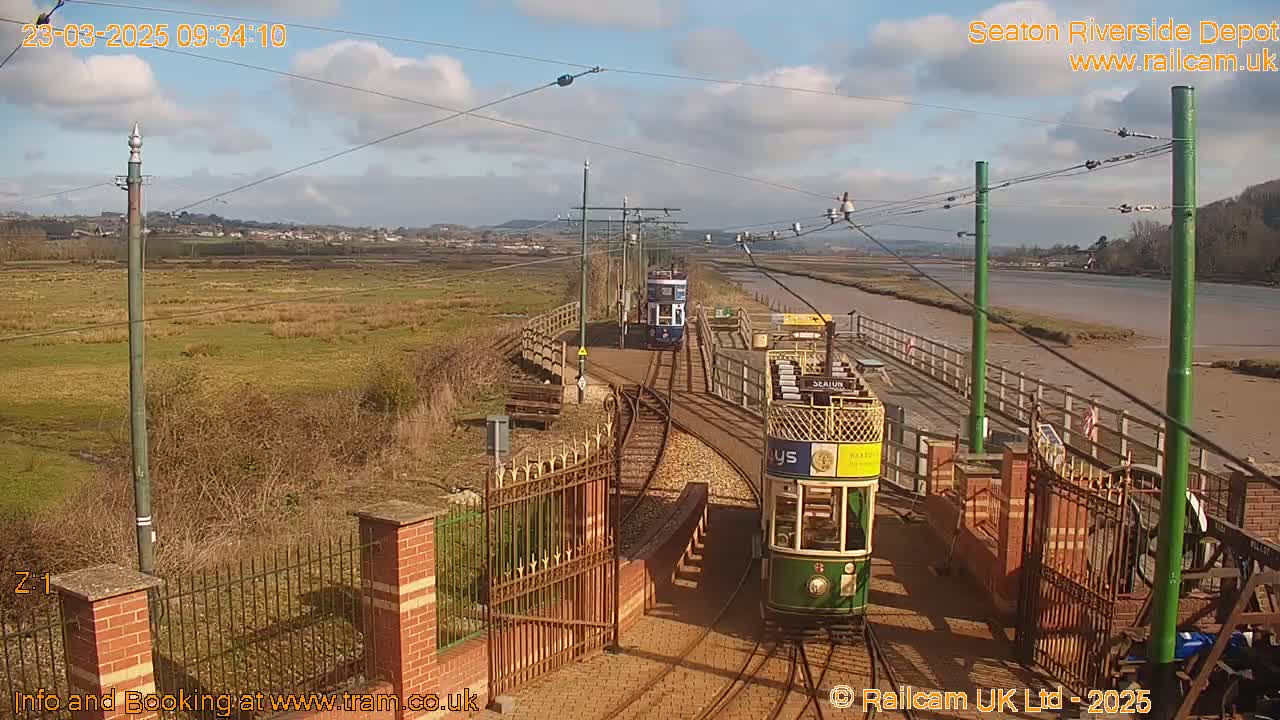 Two vintage trams are on a track near a body of water under a partly sunny sky.