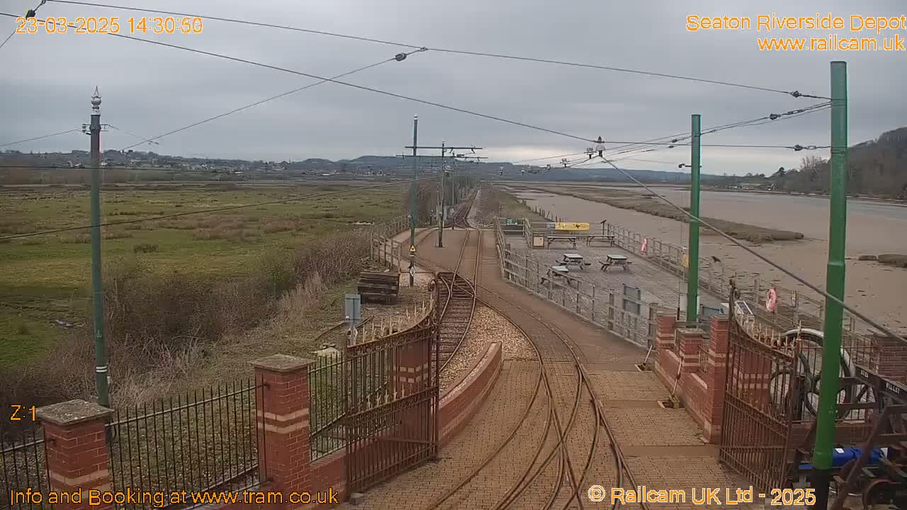 Tram tracks curve toward a waterfront depot under an overcast sky, with a muddy river visible in the background.