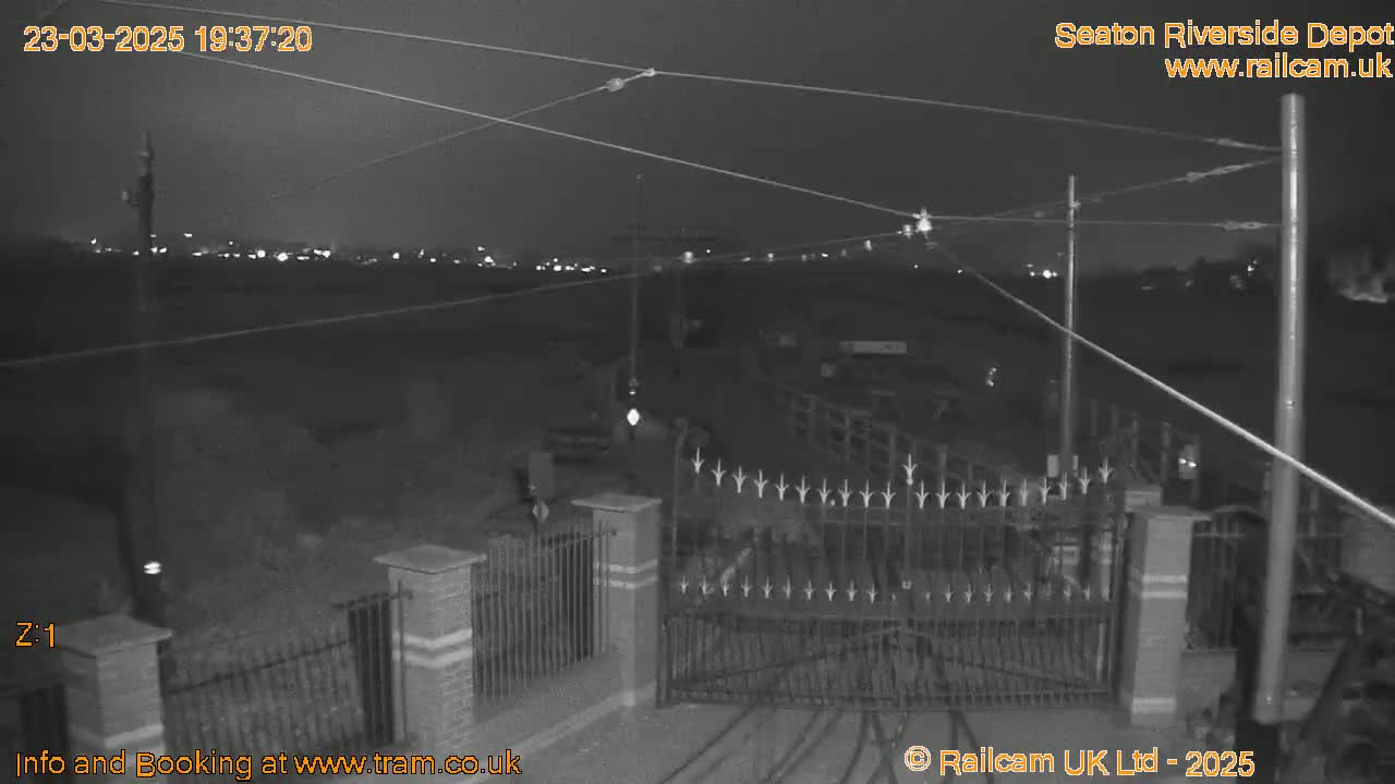 A nighttime, low-light view of a closed gate at a depot, with overhead wires and distant lights visible.
