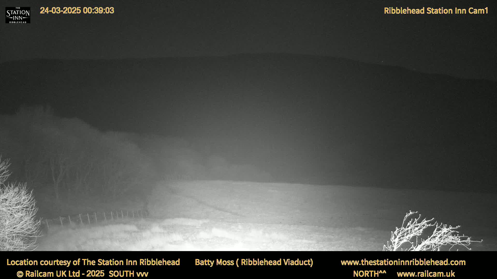 A nighttime, low-light view of a flat, snow-covered field, extending to dark hills in the distance under a dark, overcast sky.