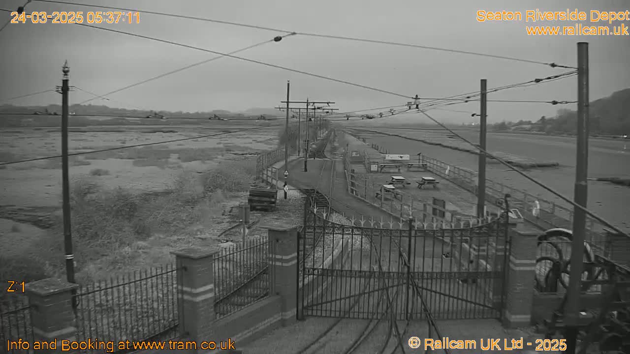 A grayscale view shows a railway depot with overhead wires, a fenced-in area with picnic tables, and overcast skies.