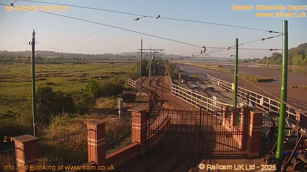 A train depot with tracks and a wooden platform sits beside a calm, muddy river under a clear, bright sky.