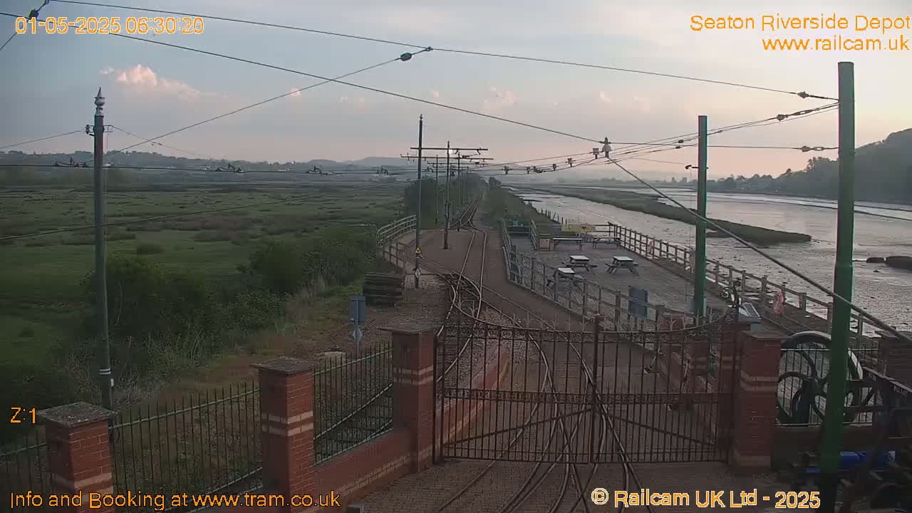 A railway depot is situated beside a calm river under a partly cloudy, dawn sky.