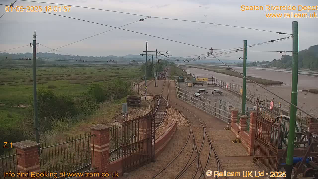 A train depot with several sets of tracks sits beside a wide, muddy estuary under an overcast sky.