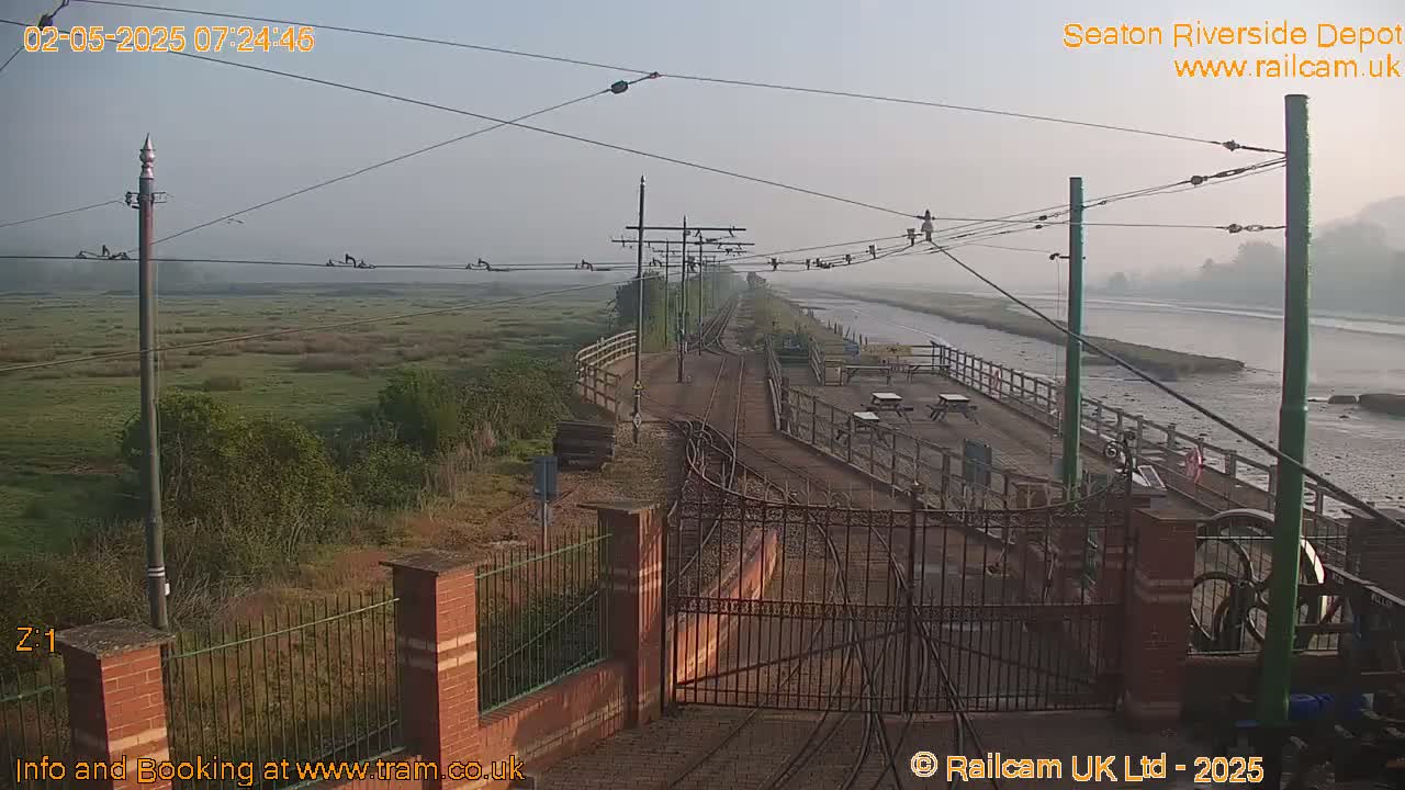 A misty morning view of a train depot alongside a river, featuring tracks, a gated entrance, and outdoor seating areas.