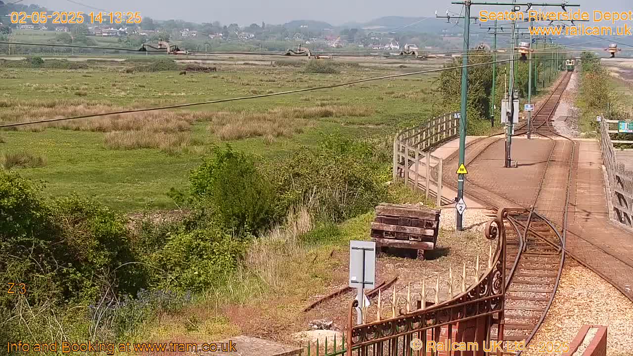 A high-angle view of a railway depot on a sunny day, showing train tracks, a grassy area, and some buildings in the distance.