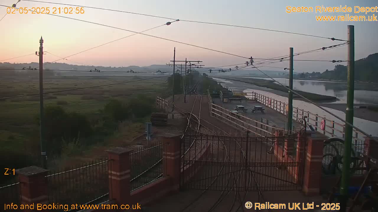 A twilight view of train tracks and a depot beside a calm river, with a grassy area and hills in the background.