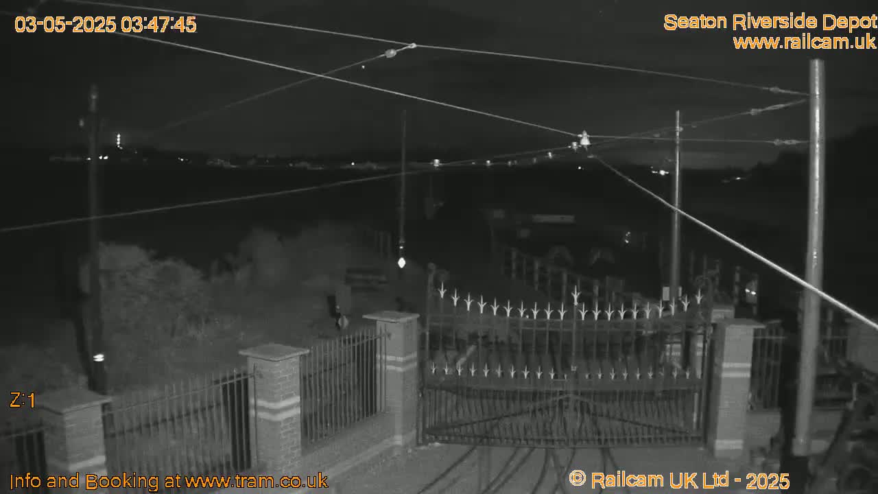 A nighttime black-and-white view shows an open metal gate at a depot, with overhead wires and a fence in the foreground.