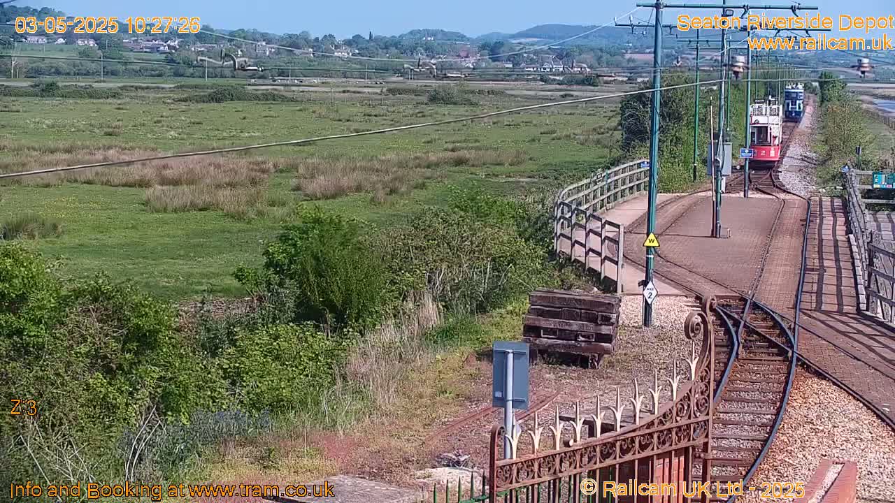 A red tram travels along railway tracks toward a depot on a sunny day, passing by a grassy area and a wooden fence.