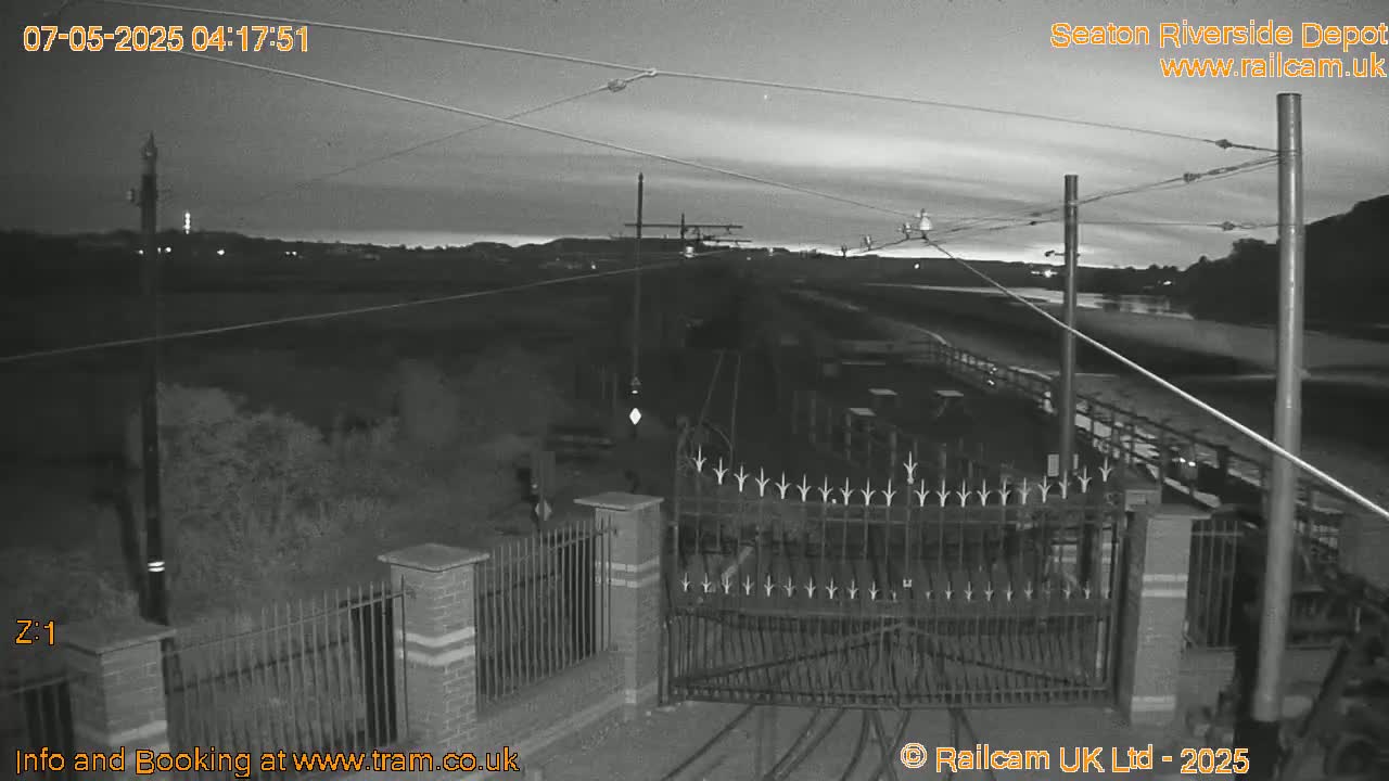 A nighttime, grayscale view of a gated entrance to a depot, with overhead wires and a body of water visible in the distance under a dusky sky.