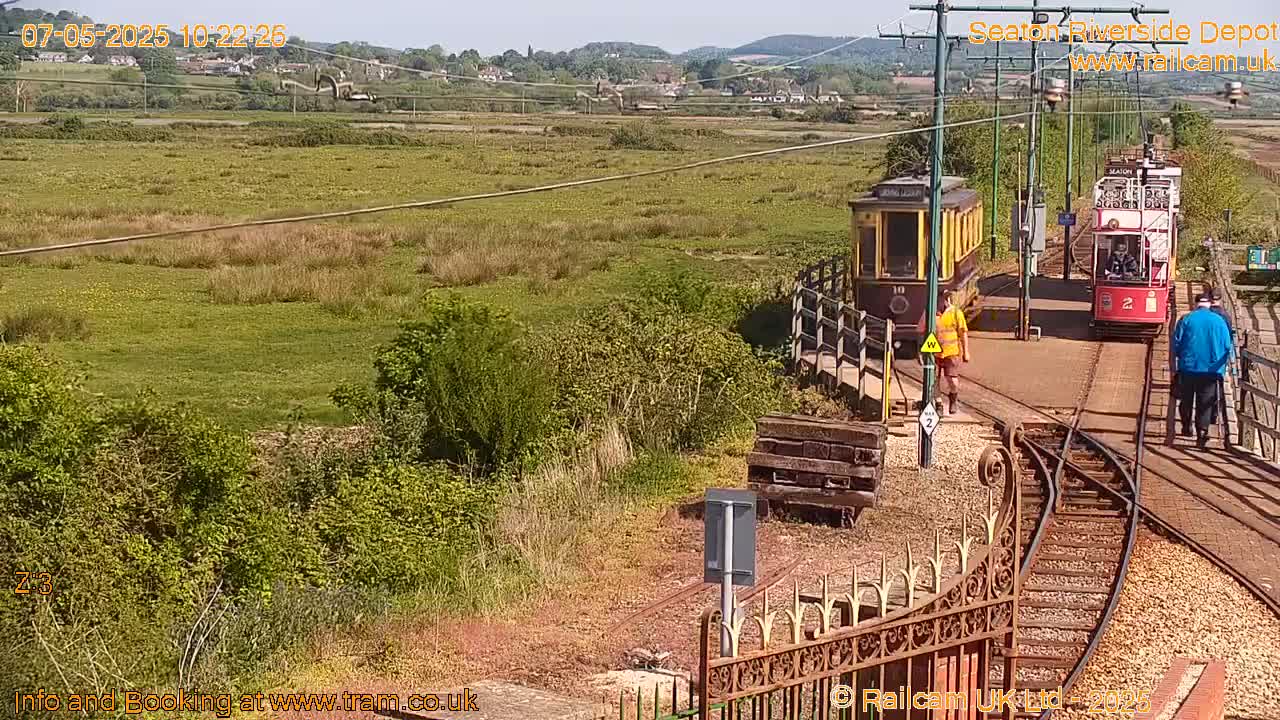 Two vintage trams are on train tracks in a sunny, grassy area with a person standing near each tram and another walking away.