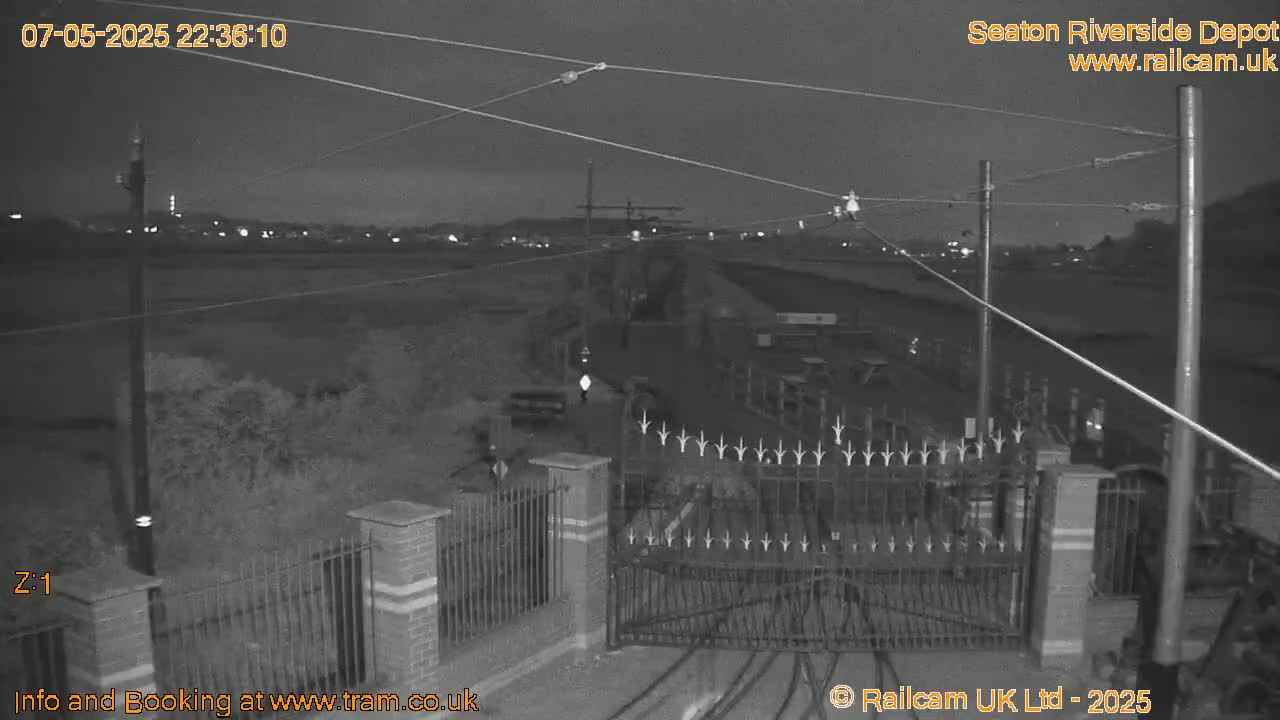 A nighttime, low-light view of a gated entrance to a depot, with overhead wires and a flat landscape extending into the background.