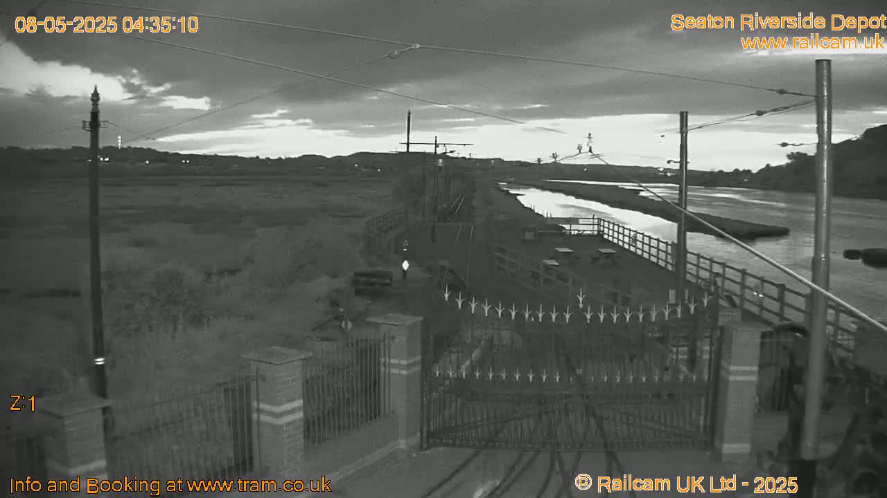 A grayscale, nighttime image shows a river, a depot area with gates and fencing, and an overcast sky.