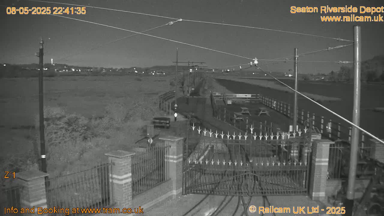 A nighttime, black-and-white view of a gated entrance to a depot, with overhead wires and a flat, open area beyond.
