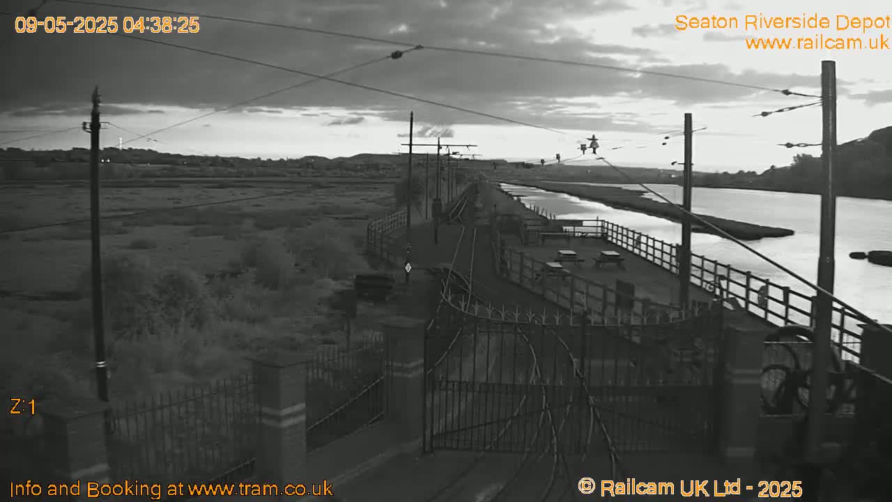 A grayscale image shows a railway line extending to a riverside depot at dawn or dusk, under a cloudy sky.
