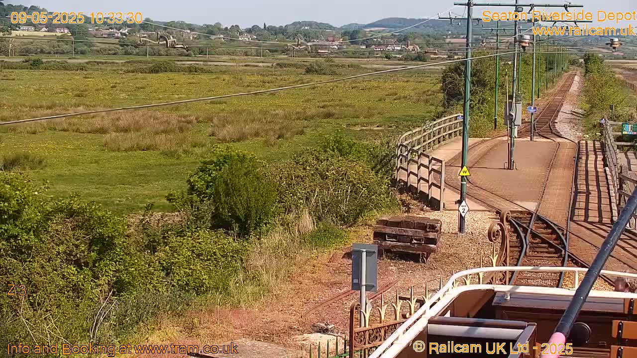 A train depot with railway tracks and a grassy area extends to a distant hillside under a sunny sky.