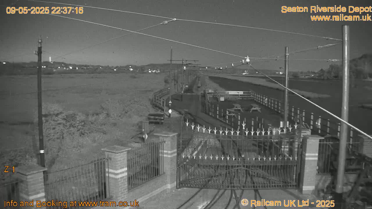 A nighttime, black-and-white view of a gated entrance to a depot, with overhead power lines and a flat, empty field beyond.