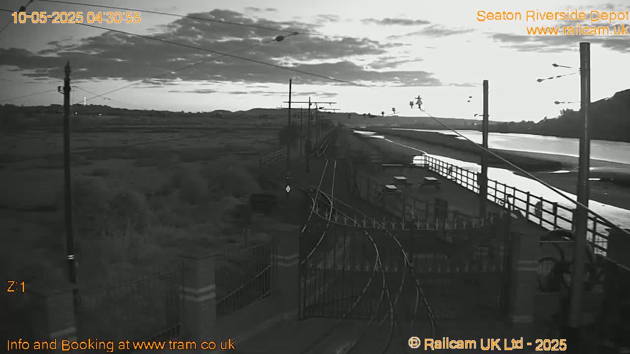 A grayscale image shows a railway depot at dawn under a cloudy sky, next to a river.