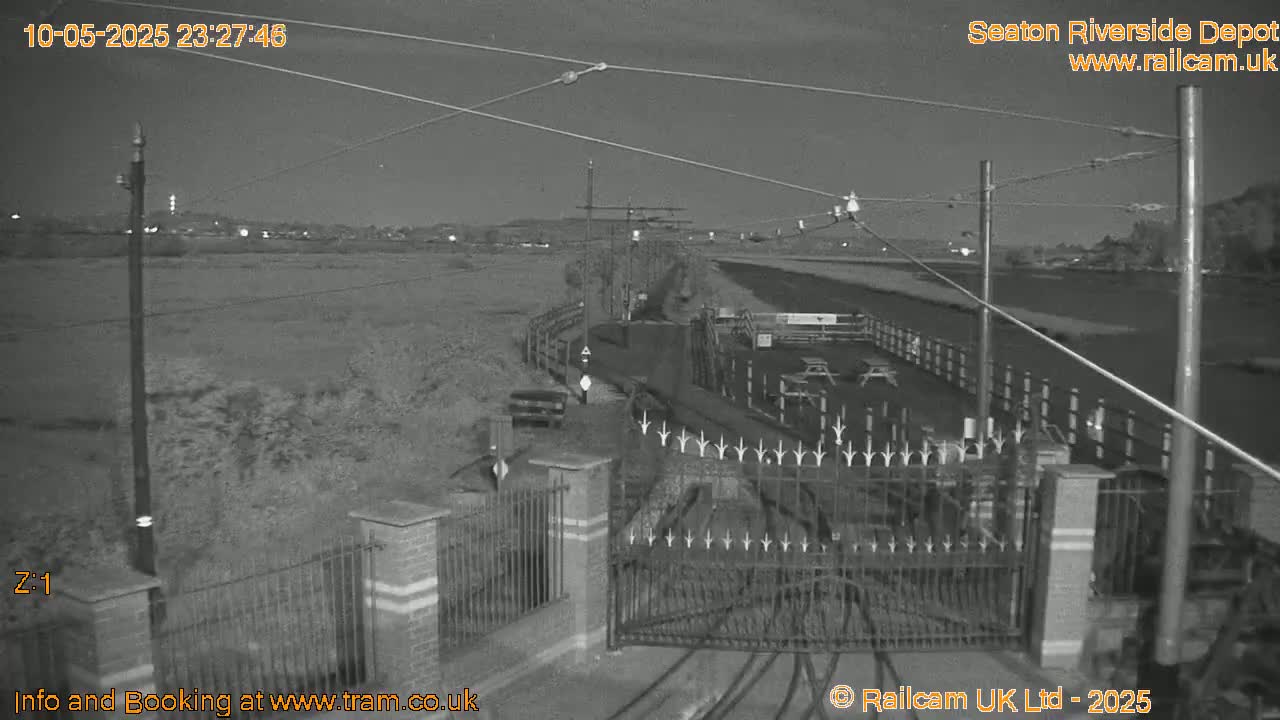 A nighttime, low-light view of a gated entrance to what appears to be a railway depot or transit area, with overhead wires and a flat, grassy area beyond.