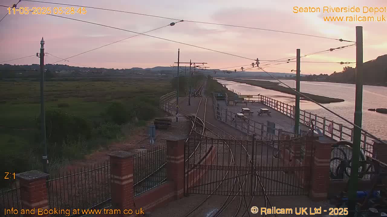 A tranquil sunrise scene shows a railway depot with tracks and gates, overlooking a calm river and marshland.