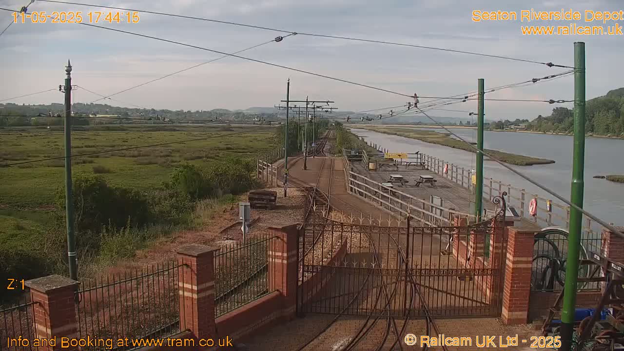 A train depot with railway tracks and a gate overlooks a calm river and marshland under a partly cloudy sky.