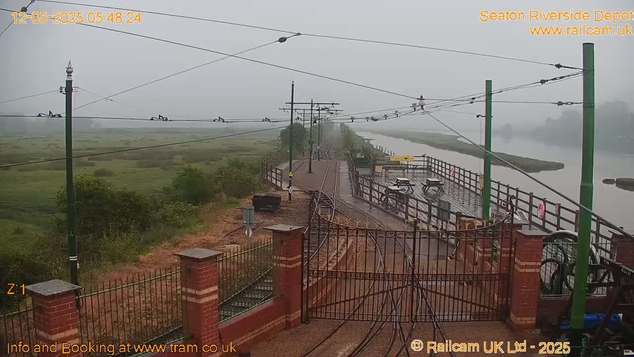 A misty view of a railway line leading to a waterfront depot with a gated entrance, alongside a river and marshland.