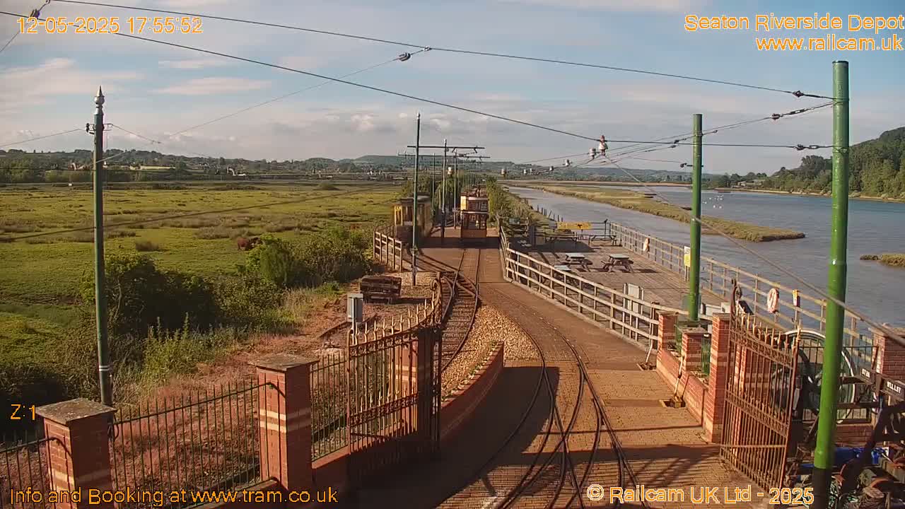A sunny day reveals a train depot along a river, featuring train tracks, a tram, and a wooden platform.