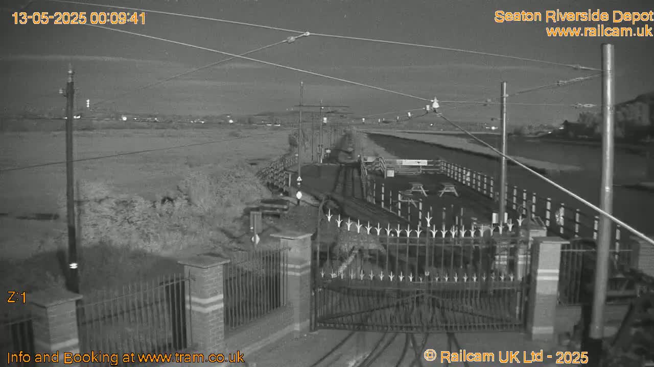 A nighttime, black-and-white view of a gated depot area next to a body of water, under a clear sky.