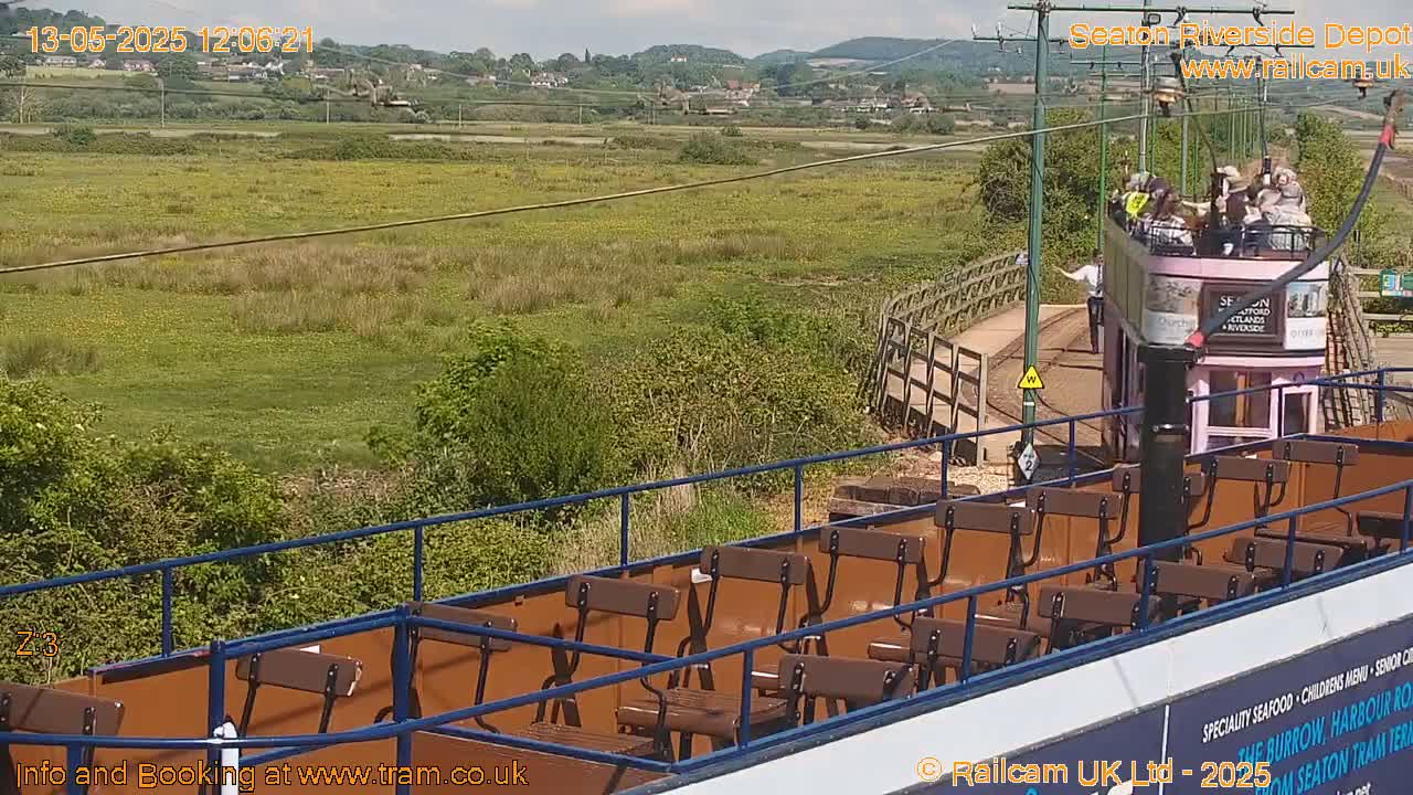 A partly sunny view from a tram shows a flat expanse of green and yellow vegetation, a small bridge, and a pink open-top tram carrying passengers.
