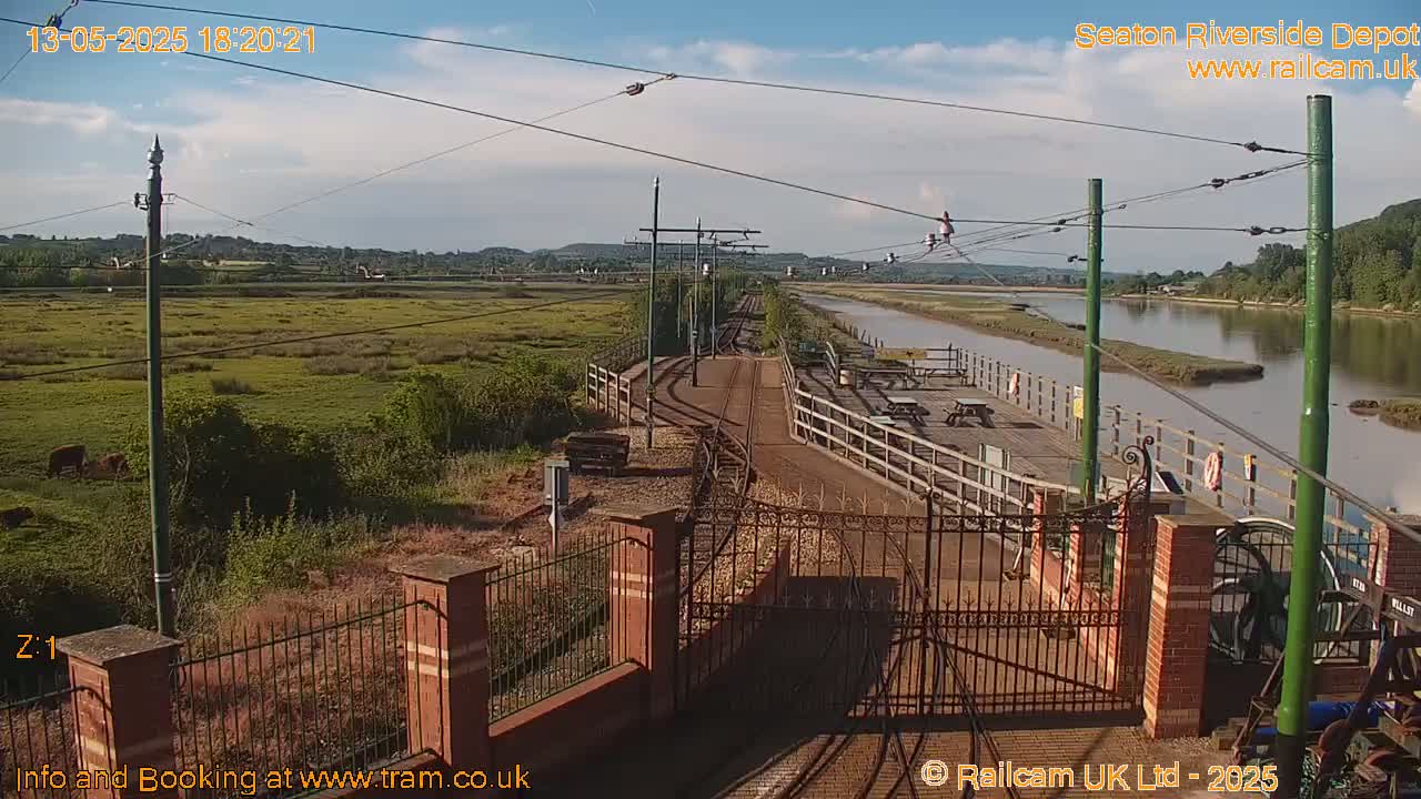 A railway depot with train tracks and a gate, overlooking a calm river and marshland under a mostly clear sky.