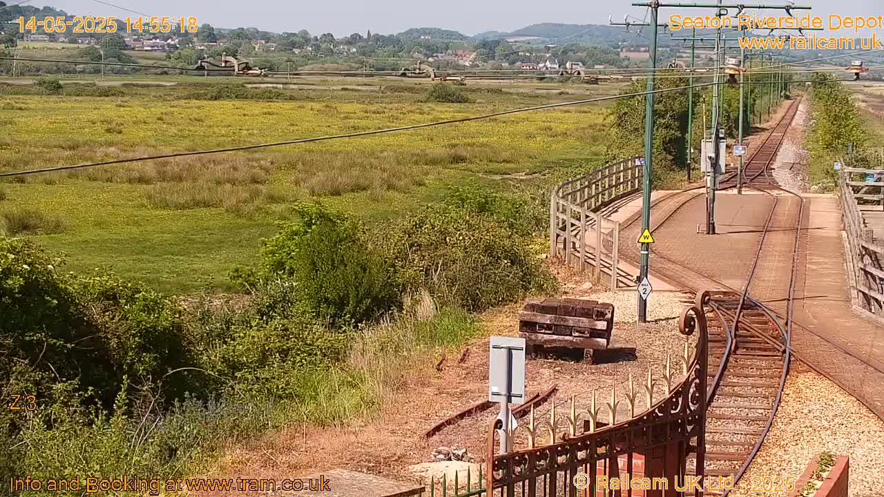 A sunny view of a train depot with railway tracks and a grassy field extending to a distant hillside.