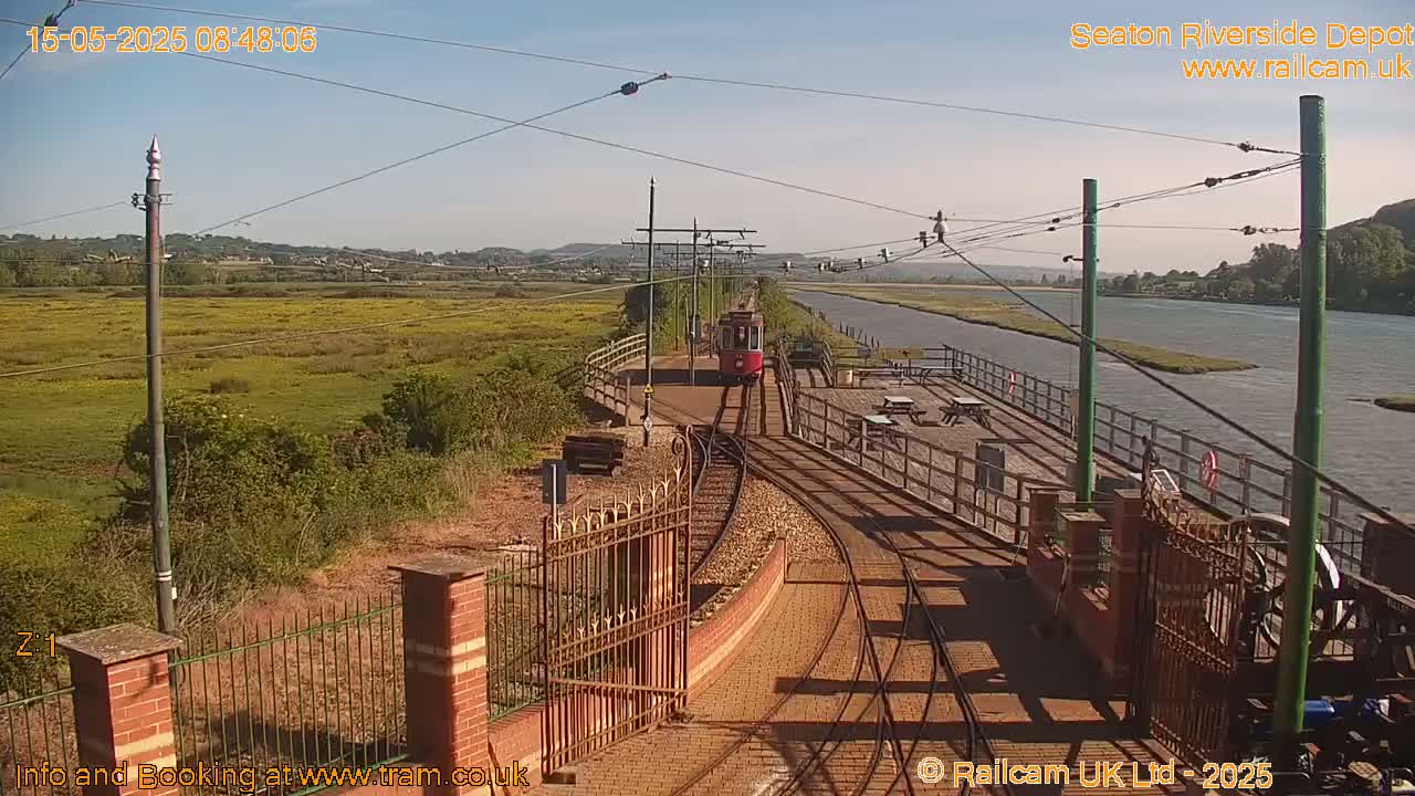 A red tram approaches a station on a sunny day next to a river and grassy wetlands.
