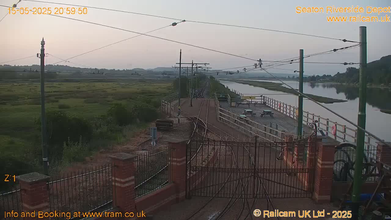 A twilight view of a railway track leading to a waterfront area with picnic tables, bordered by a metal fence and calm water.