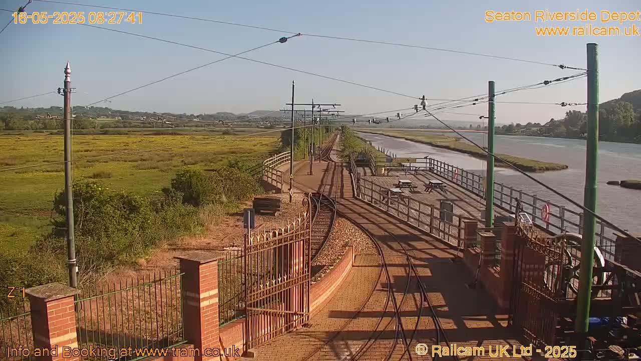A sunny view of train tracks leading to a depot beside a river and a grassy field.