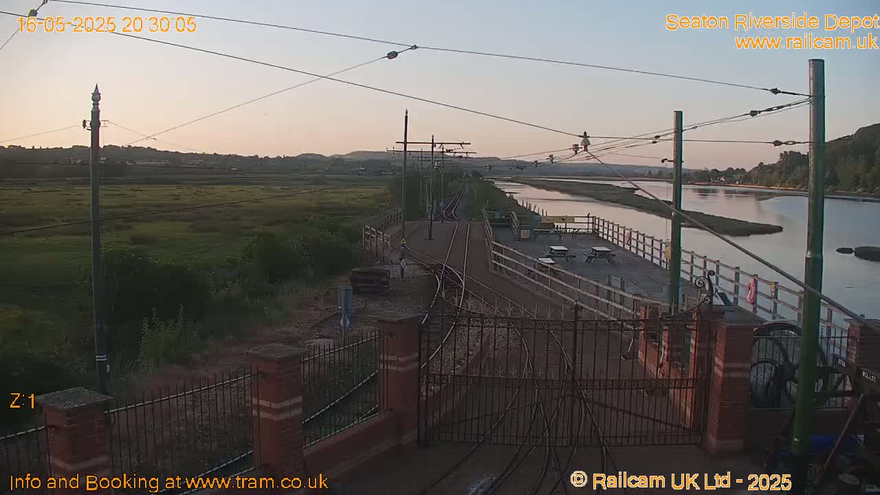 A railway track at dusk leads to a waterfront platform with picnic tables, next to calm water and marshland.