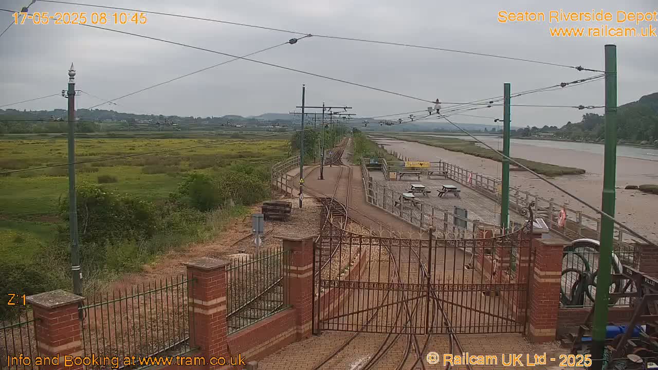 A railway depot with overhead wires, located next to a river under an overcast sky, features tracks, gates, and a fenced-in area with picnic tables.