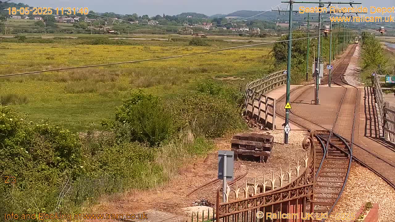 A sunny view of train tracks curving through a grassy area with yellow wildflowers, leading to a distant village and hills.