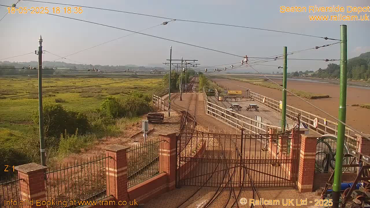 A sunny view of a railway depot with tracks, a gate, and a wooden platform extending towards a muddy, low-tide shoreline beyond a grassy field.